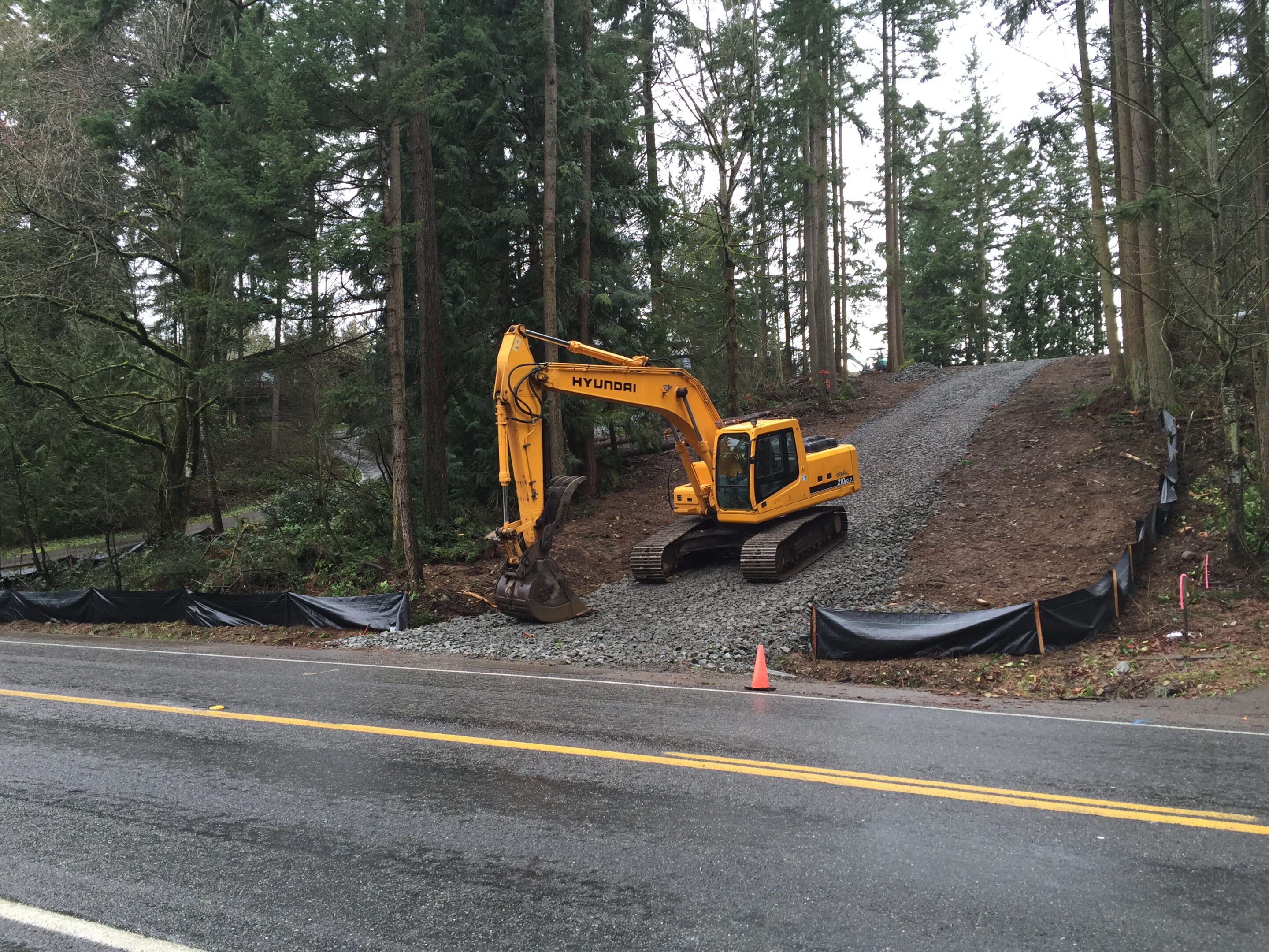 Yellow excavator digging near a forest by the roadside.