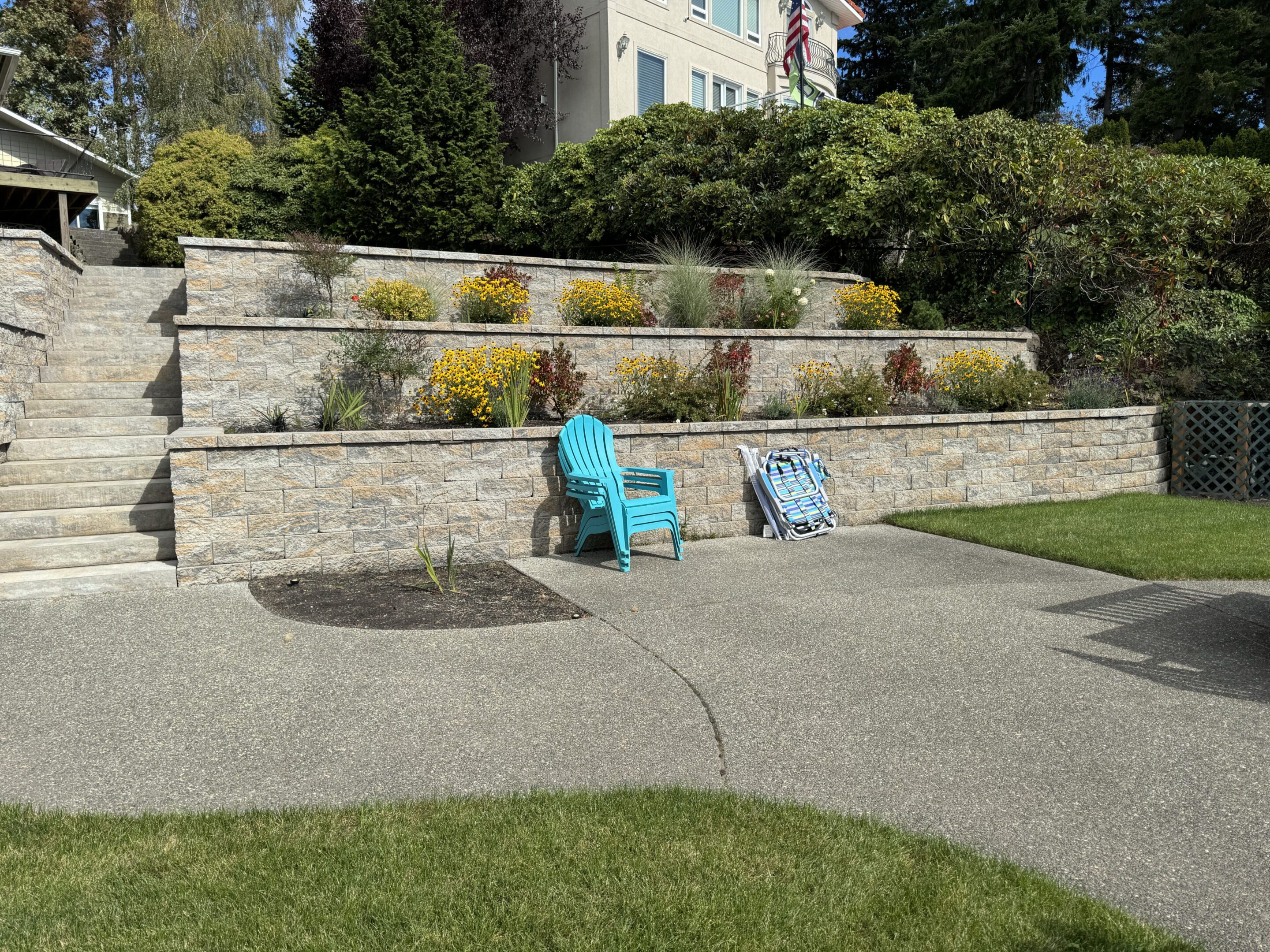 Two chairs and an umbrella resting against a stone retaining wall in a sunny garden.